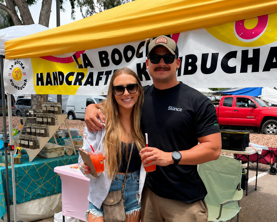 A man and a woman posing in front of a yellow and white canopy at an outdoor market. The canopy features the text 'LA BOUCHERIE HANDCRAFTED KEBUCHA' and the man is wearing a black t-shirt with 'skate' printed on the chest.