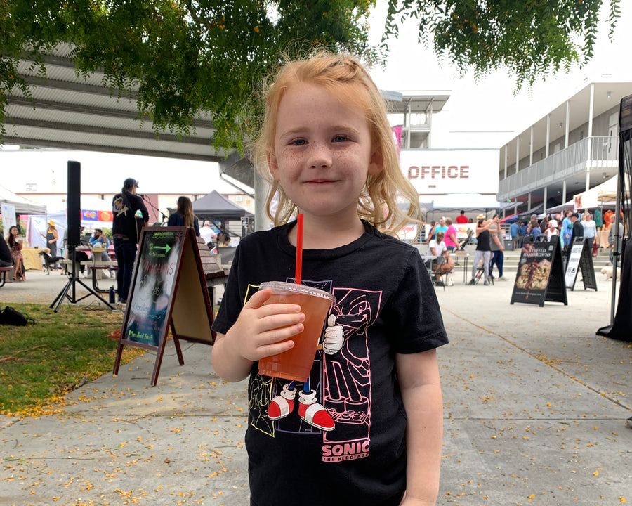 A young girl with blonde hair stands outdoors at what looks like a festival or event, wearing a black graphic t-shirt with a Sonic the Hedgehog design and pink leggings, holding an iced coffee.