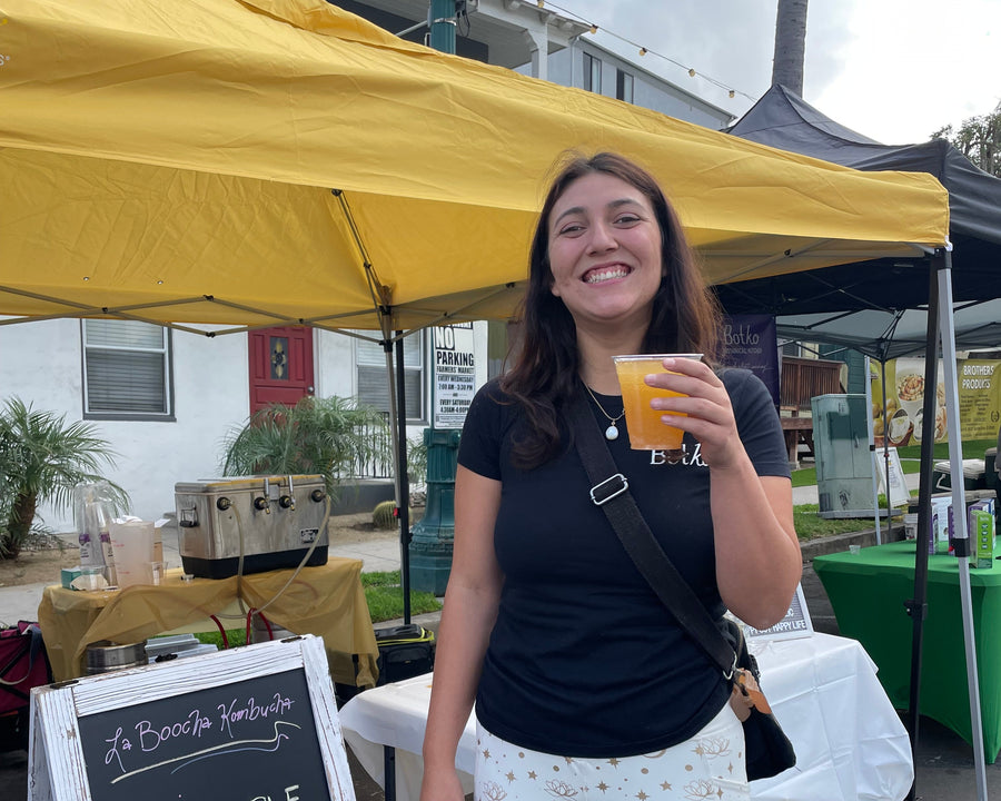 A smiling woman stands outdoors under a yellow canopy at what appears to be a street food or craft event, holding a small orange cup. A chalkboard sign in front lists flavors including Pineapple Jalapeño, Mango Tatin, and free samples.