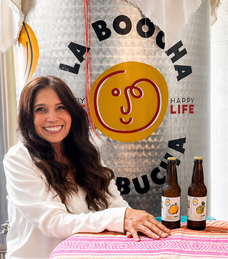 A smiling woman in a white blouse stands in front of a large stainless steel vat with a white cloth drape and the visible brand name La Boocha. Two brown beverage bottles sit on a pink patterned tablecloth in the foreground.
